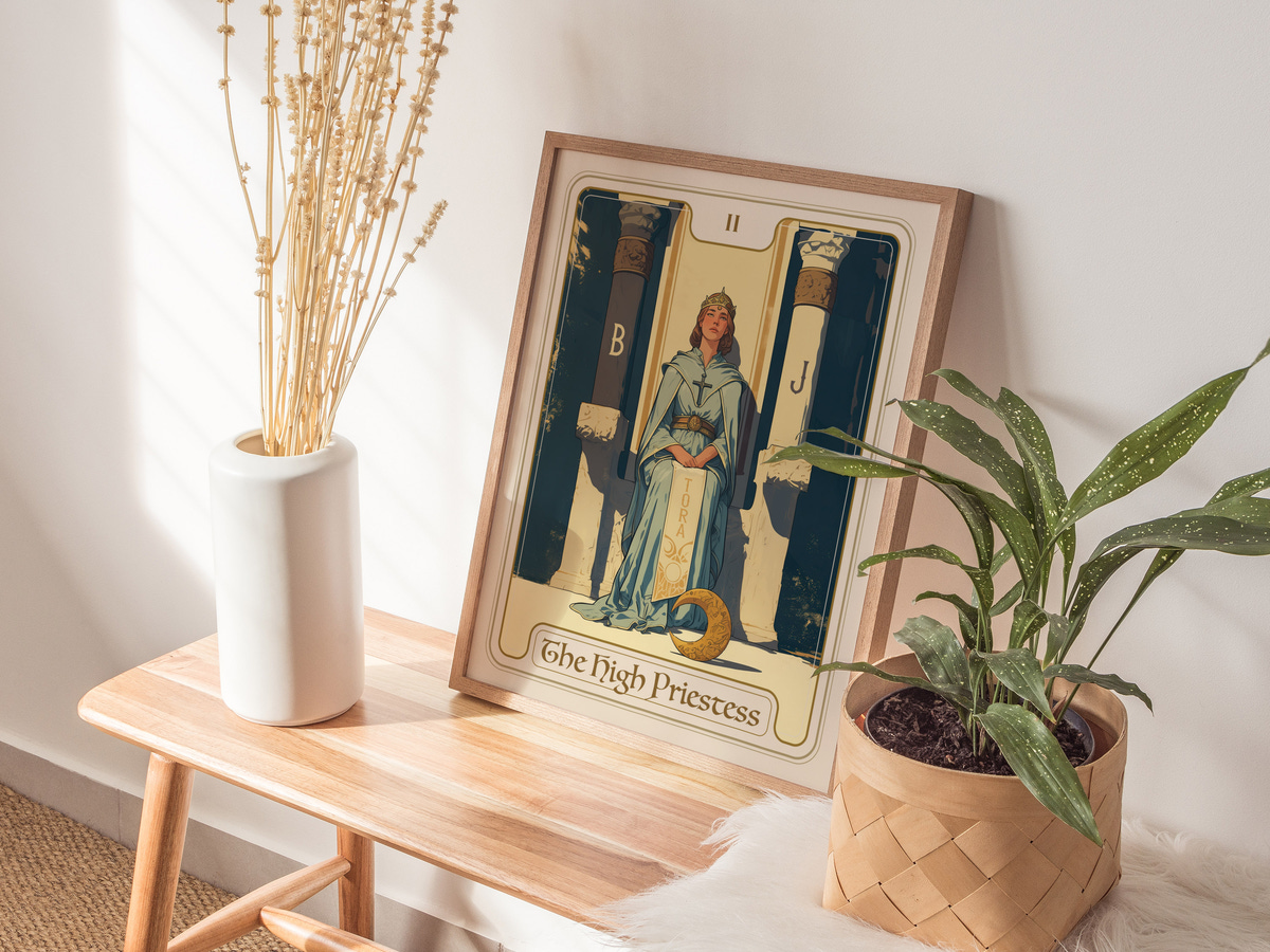 A framed art print titled The High Priestess is displayed on a wooden table next to a white vase containing dried flowers and a green potted plant.