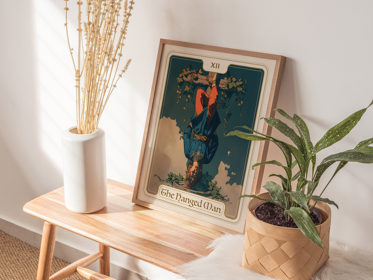 A framed art print of The Hanged Man tarot card is displayed on a wooden table, featuring a figure hanging upside down amidst greenery. Nearby, a tall white vase holds dried flowers, and a potted plant adds a touch of greenery to the scene.