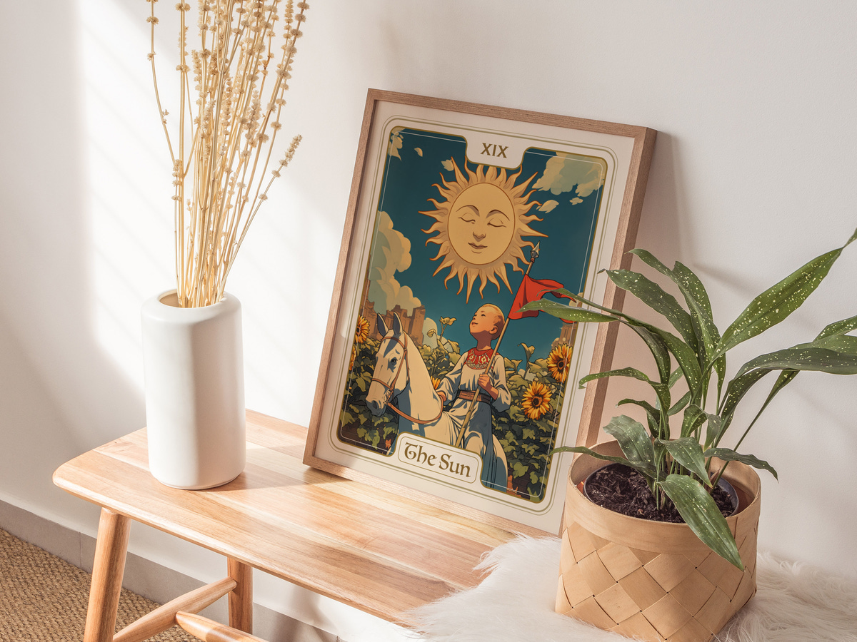A framed art print titled The Sun features a whimsical sun illustration with a smiling face, surrounded by vibrant flowers. The artwork is displayed on a wooden table alongside a white vase containing dried flowers and a potted plant.