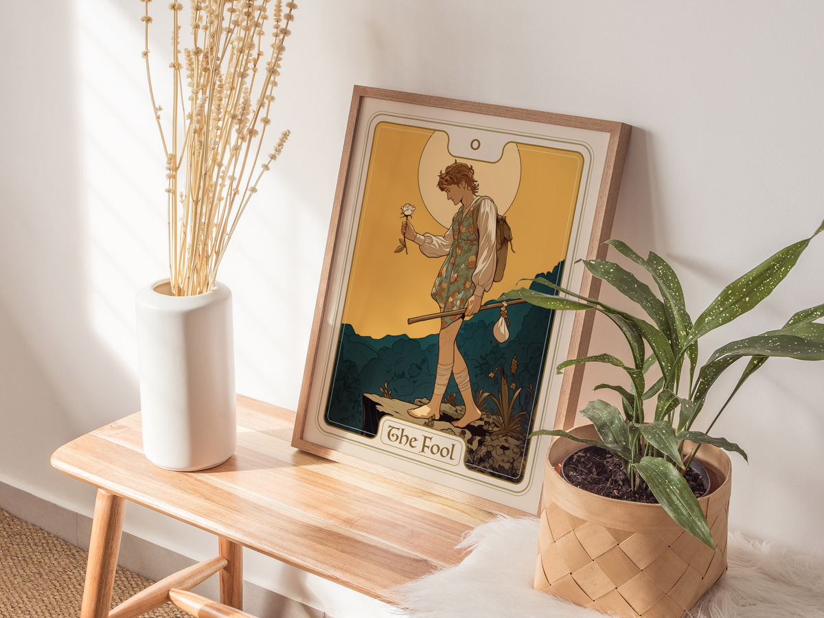 A stylish framed art print titled The Fool is displayed on a wooden table, featuring a whimsical illustration of a young person holding a flower against a vibrant yellow background.