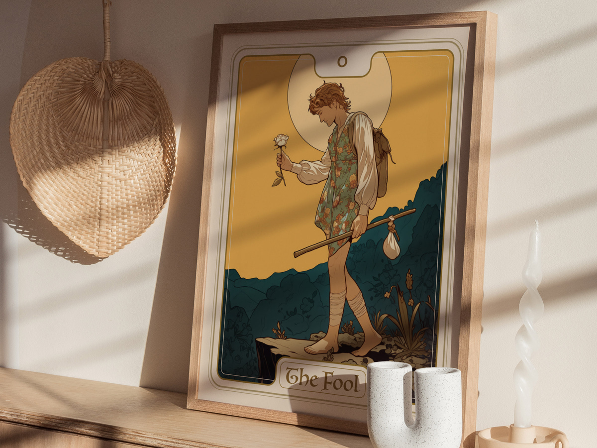A framed illustration titled The Fool features a young man standing on a rocky path, holding a flower and carrying a stick with a small bag. The background showcases a warm yellow sky and lush greenery, creating a whimsical atmosphere.