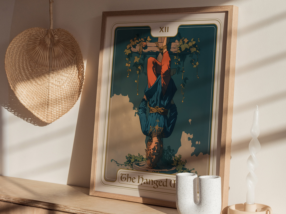 A framed art print titled The Hanged Man features an illustration of a man hanging upside down, surrounded by greenery. The artwork is set against a blue background and is displayed on a wooden shelf beside decorative items.