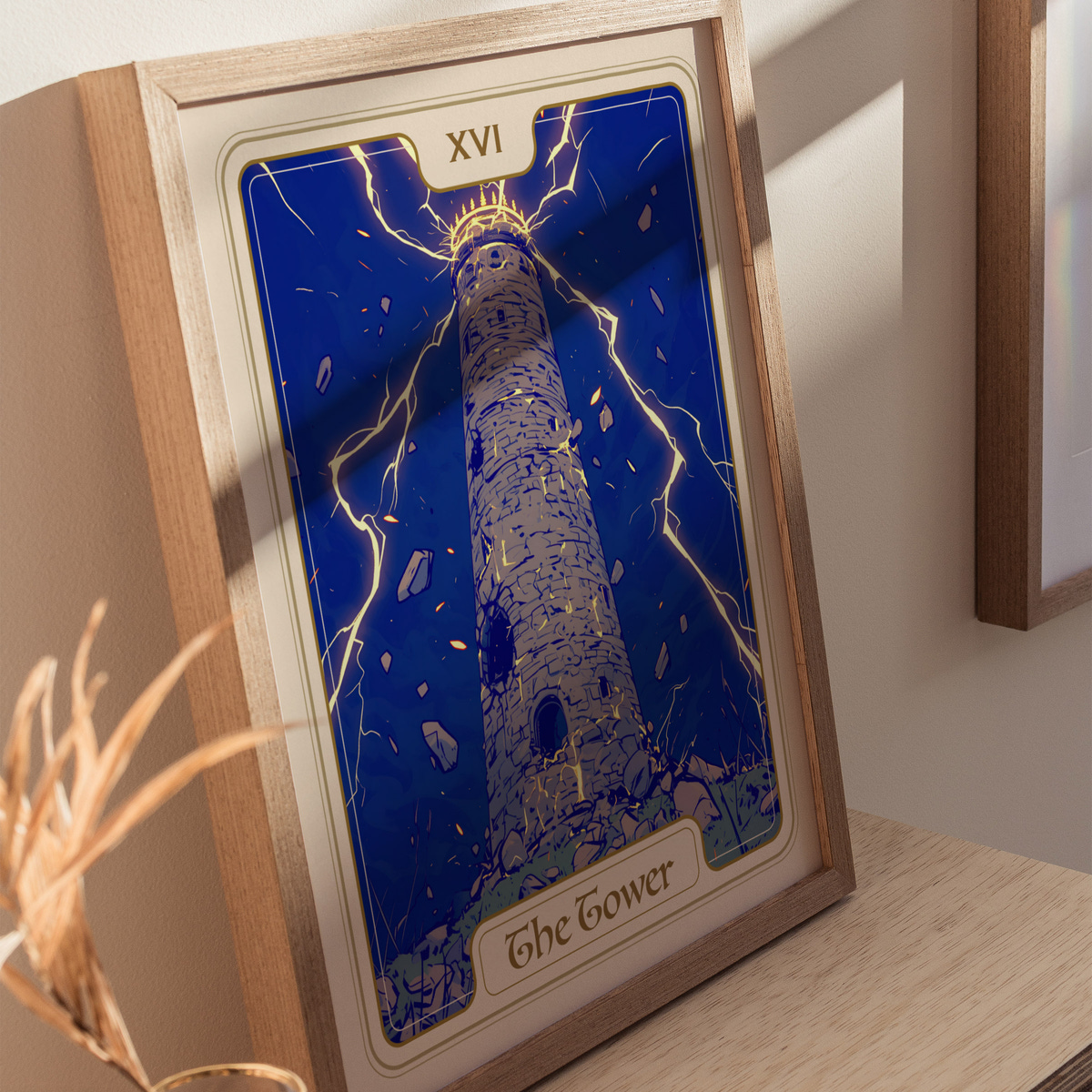 A framed illustration of The Tower, featuring a tall, stone tower surrounded by dramatic lightning against a deep blue sky. The artwork is displayed on a light-colored wooden shelf, with a plant in the foreground.