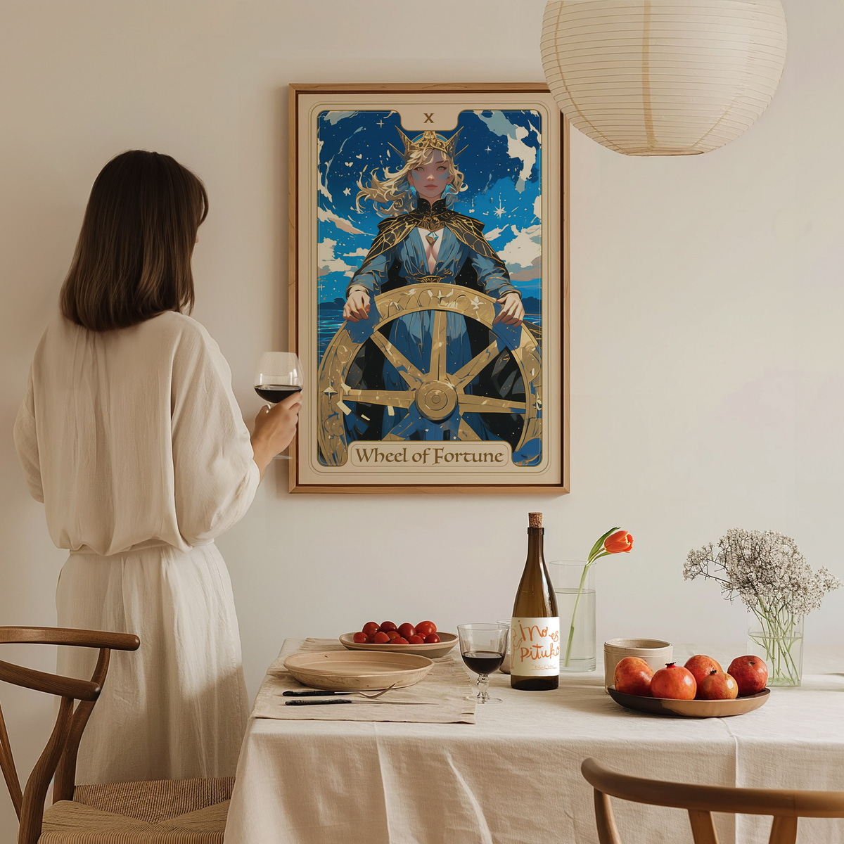 A woman in a white outfit stands in a softly lit room, gazing at a framed poster titled Wheel of Fortune, which features a regal figure at a ships wheel under a blue sky. A dining table is set with a bottle, glasses, and fresh fruit.