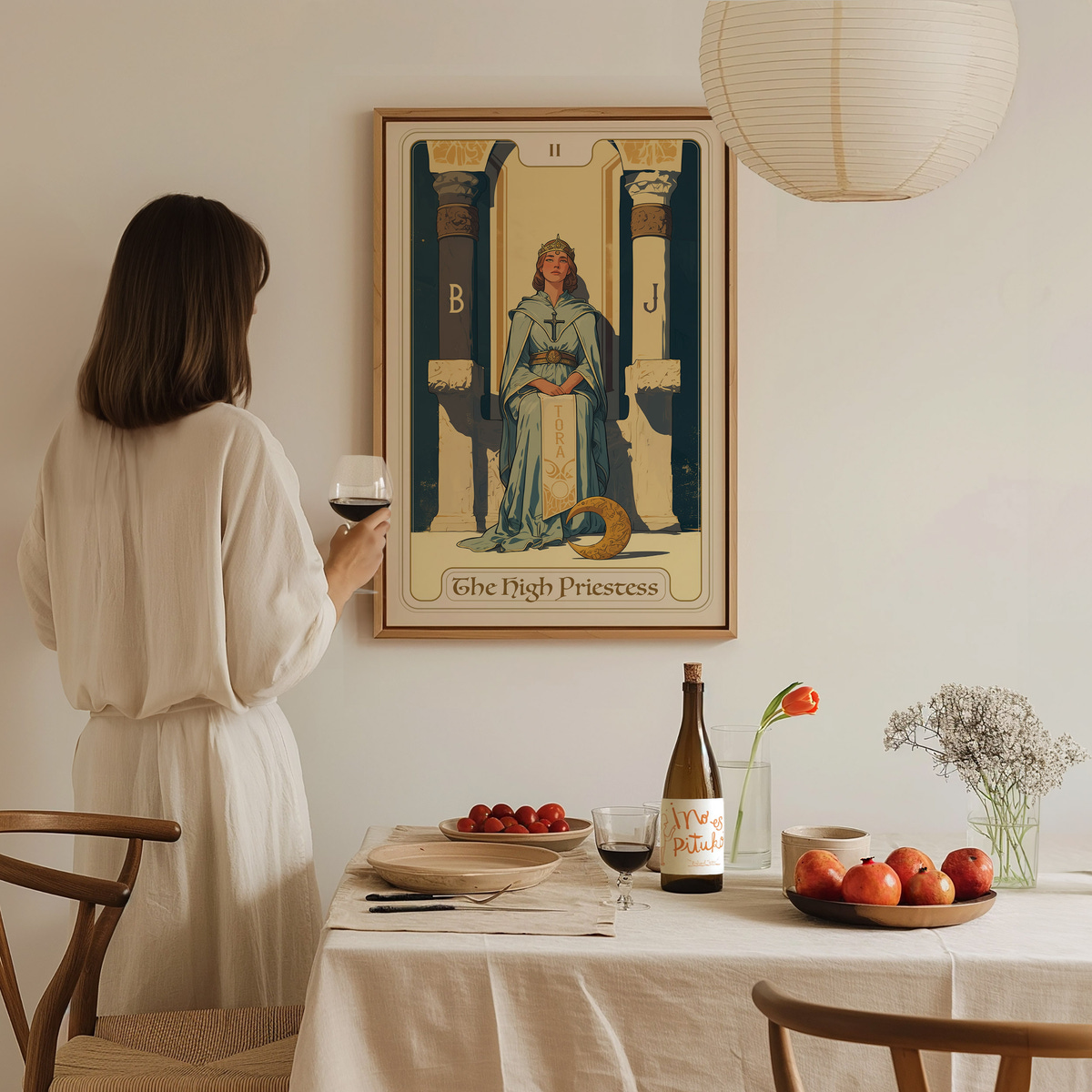 A woman in a white dress stands with a glass of red wine, gazing at a framed artwork titled The High Priestess on a light-colored wall.