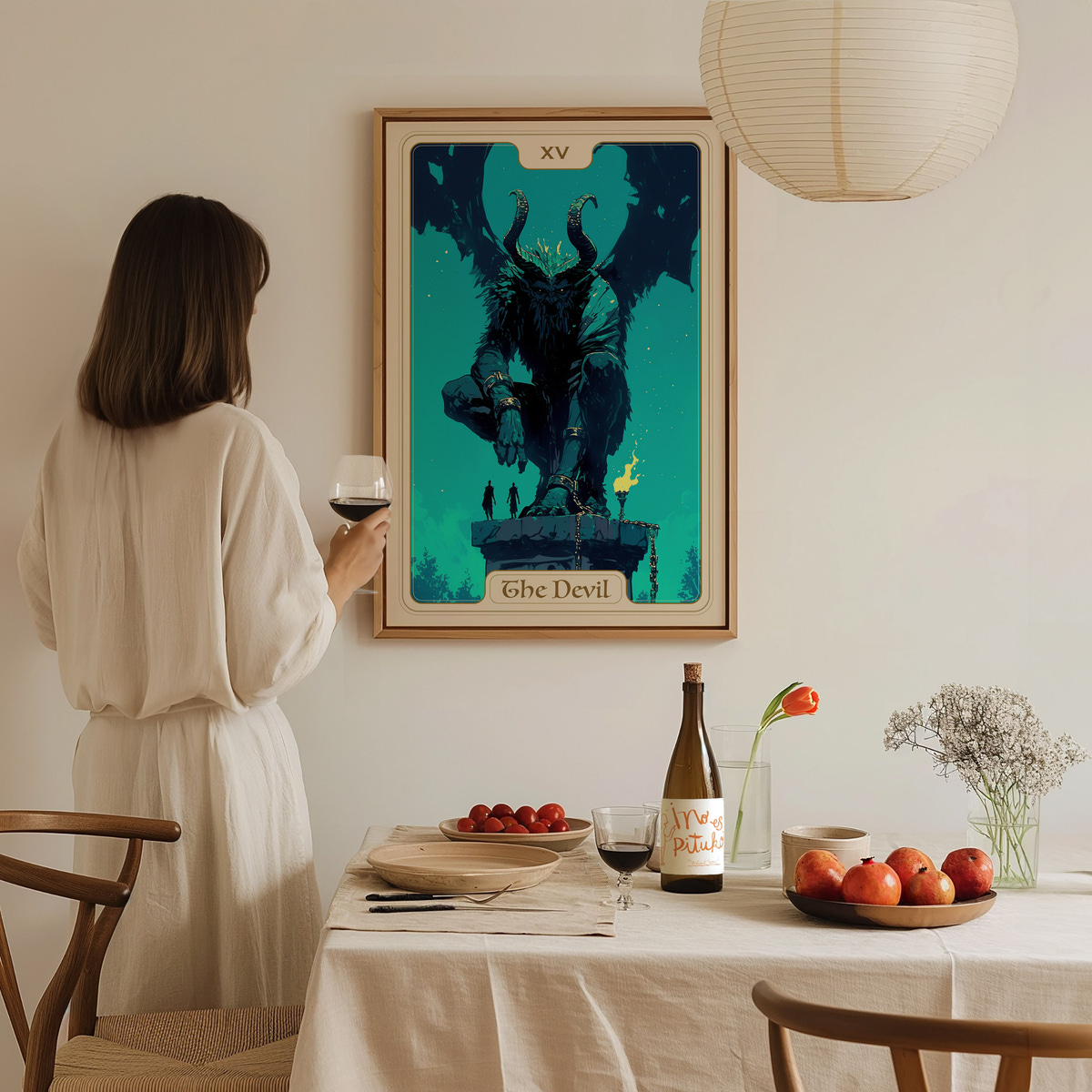 A woman in a light-colored dress stands at a dining table holding a glass of wine, gazing at a large, framed tarot card titled The Devil.
