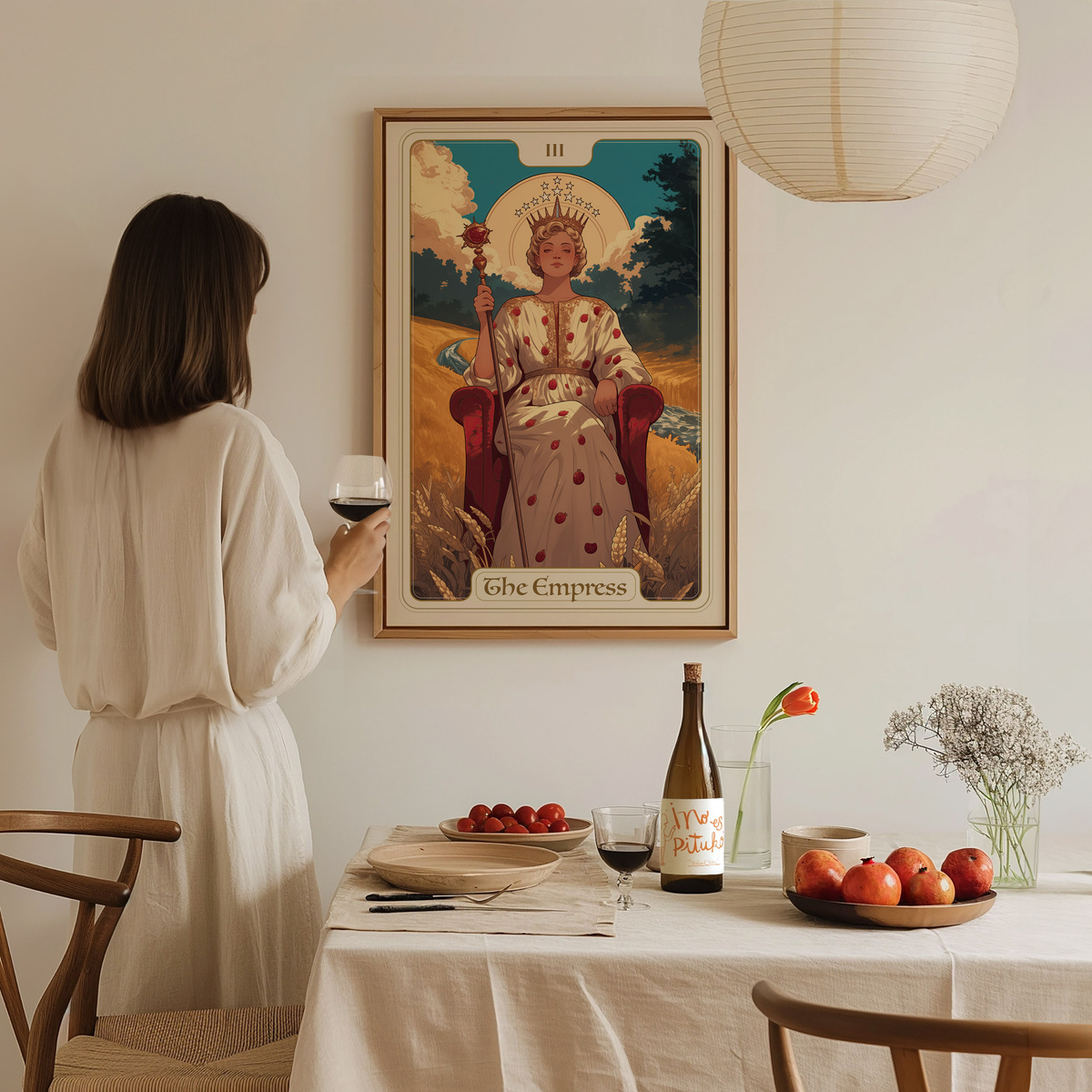 A woman in a white dress stands in a cozy dining area, holding a glass of wine, gazing at a framed art piece titled The Empress.
