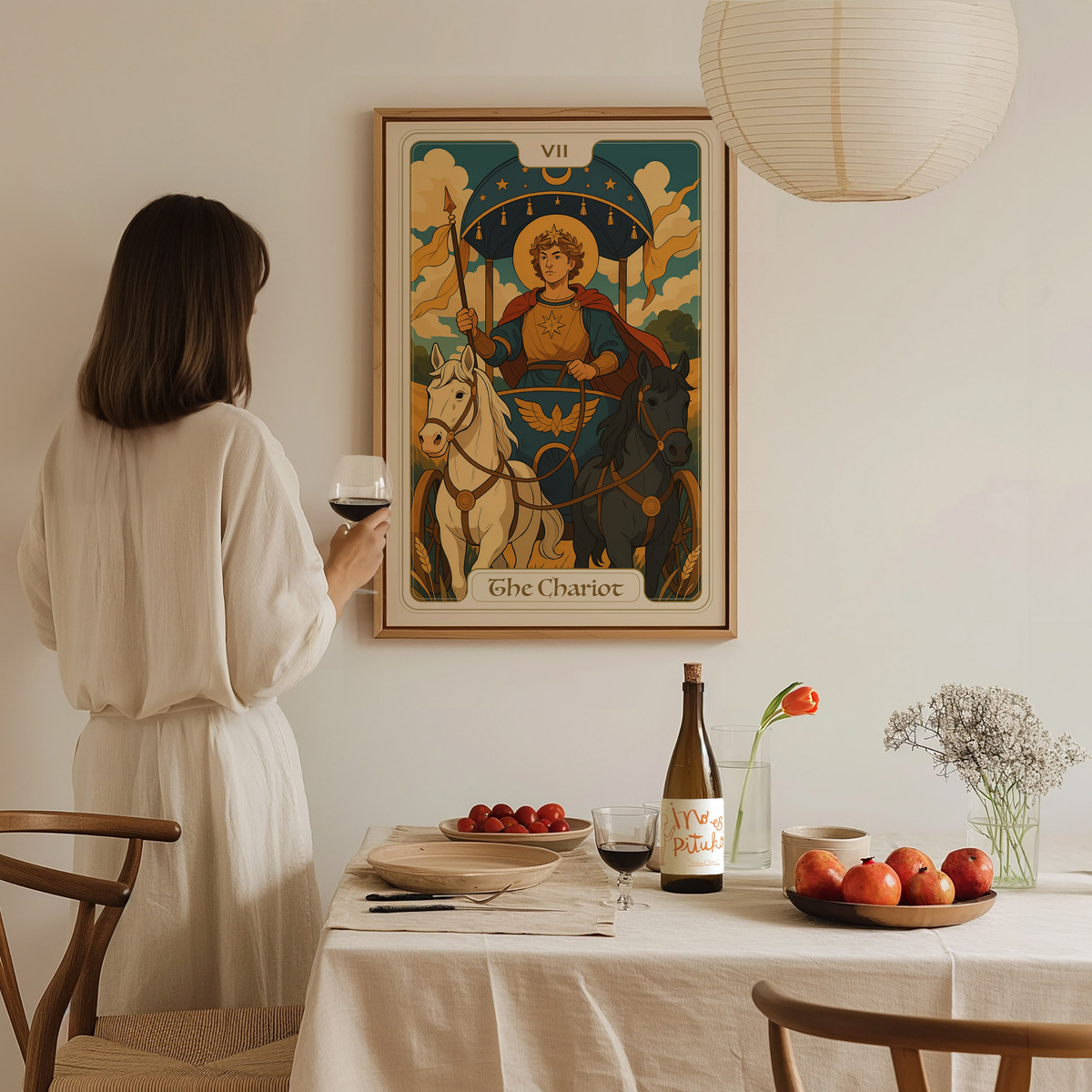 A woman in a white dress stands at a dining table, holding a glass of red wine, gazing at a colorful framed artwork titled The Chariot depicting a figure with a starry backdrop, flanked by two horses.