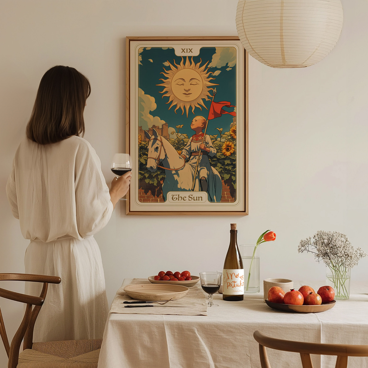 A woman in a light-colored dress stands with a glass of wine, gazing at a vintage-style poster titled The Sun, depicting a sun with a face and a figure on a horse. The cozy dining table is set with fruits, a bottle, and a vase of flowers.