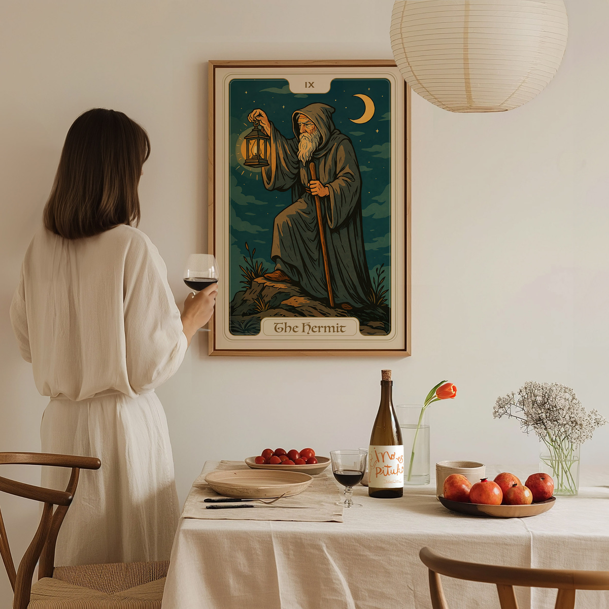 A woman wearing a light-colored outfit stands in a cozy dining area, holding a glass of wine and gazing at a framed tarot card titled The Hermit on the wall.