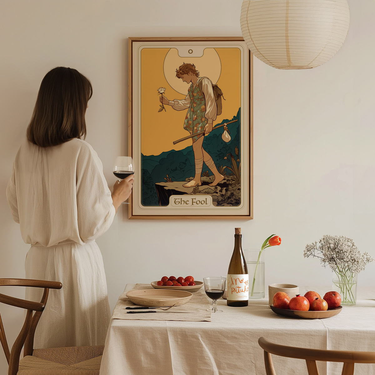 A woman in a light-colored dress stands at a dining table, holding a glass of wine and gazing at a framed art piece titled The Fool on the wall.